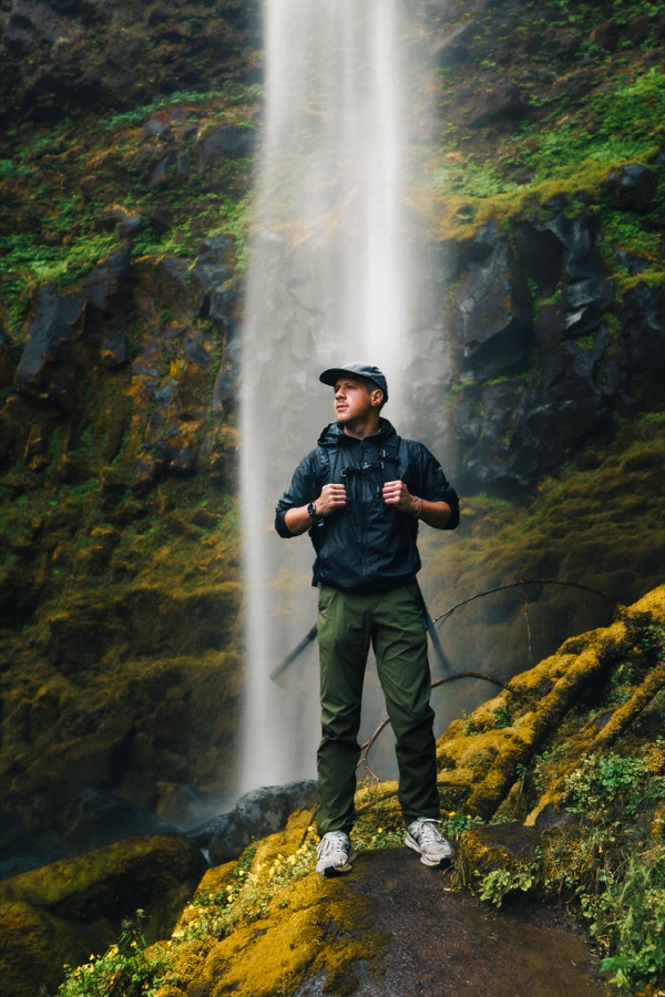 Logan standing on a mossy ledge at the foot of a tall waterfall, in a cap and rain shell with his pack straps in both hands
